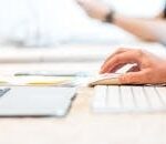 A person working at a laptop with a wireless mouse and keyboard in a bright office setting.