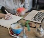 Woman writing in a notebook with a laptop and coffee cup on a desk. Ideal for workspace inspiration.