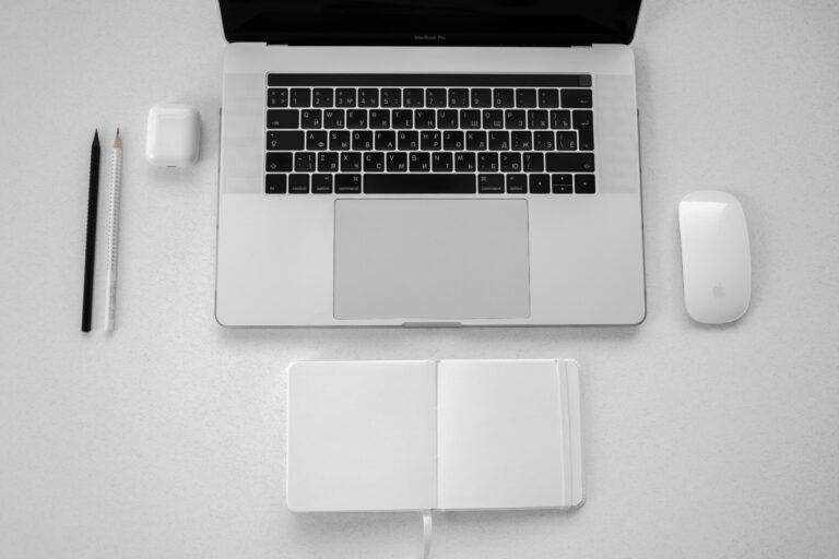 A minimalist top-down view of a workspace featuring a laptop, notebook, pencils, and wireless mouse.